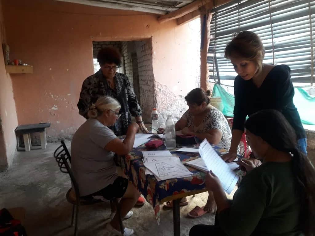Taller de alfabetización para adultos: grupo de mujeres alrededor de una mesa con material educativo y una facilitadora en la pizarra.